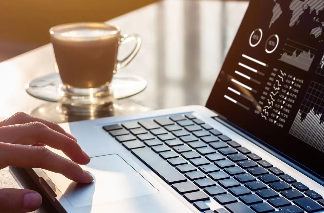 Person working on laptop computer with business analytics and financial report data with metrics and charts on screen at modern office workplace with sunset light and coffee